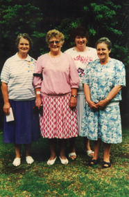 Four older women stand side by side smiling with trees and shrubbery behind them. 