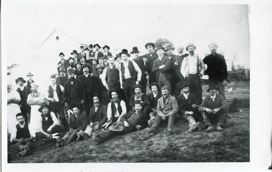 Black and white photograph of a large group of men, most with moustaches and wearing hats, stand together in front of a gabled building (Allen's or Kerferd's brewery?). Some of the men are seated or reclining on the grassy crest of the hill, and two seated men and one standing carry large shears/clippers in their right hands. On the left of the photo, two standing men wear aprons.