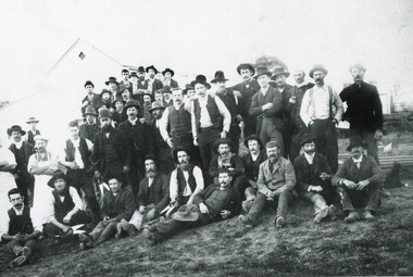 Black and white photograph of a large group of men, most with moustaches and wearing hats, stand together in front of a gabled building (Allen's or Kerferd's brewery?). Some of the men are seated or reclining on the grassy crest of the hill, two seated and one standing men carry large shears/clippers in their right hands. On the left of the photo, two standing men wear aprons.
