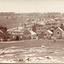 Sepia photograph of a row of business buildings on a rocky, sloping hillside, with a view across to residences and open land. Businesses from left to right (up the slope): J.E. Bishop, Coach Builders; Straughair Duncan, Engineers, Blacksmiths & Farriers; Straughair Duncan, Beechworth Foundry;  Wholesale & Retail Est. 1853?, Family Store Mackenzie; T. Pratten, Grocer - ?? & ?? Dealer. Eleven men may be seen outside the Foundry building. This is Bridge Road, Newtown, Beechworth.