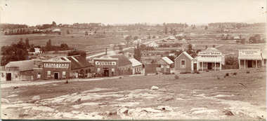 Sepia photograph of a row of business buildings on a rocky, sloping hillside, with a view across to residences and open land. Businesses from left to right (up the slope): J.E. Bishop, Coach Builders; Straughair Duncan, Engineers, Blacksmiths & Farriers; Straughair Duncan, Beechworth Foundry;  Wholesale & Retail Est. 1853?, Family Store Mackenzie; T. Pratten, Grocer - ?? & ?? Dealer. Eleven men may be seen outside the Foundry building. This is Bridge Road, Newtown, Beechworth.