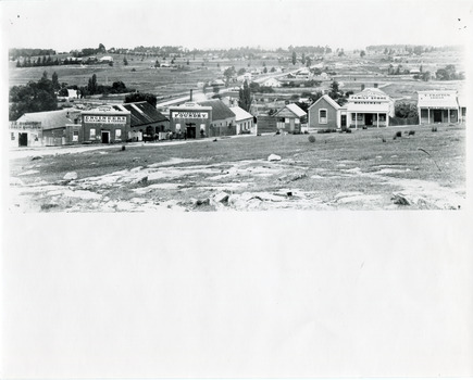 Uncropped black and white photograph (top half of paper) of a row of business buildings on a rocky, sloping hillside, with a view across to residences and open land. Businesses from left to right (up the slope): J.E. Bishop, Coach Builders; Straughair Duncan, Engineers, Blacksmiths & Farriers; Straughair Duncan, Beechworth Foundry;  Wholesale & Retail Est. 1855 (?), Family Store Mackenzie; T. Pratten, Grocer - ?? & ?? Dealer, 