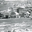 Black and white photograph of a row of business buildings on a rocky, sloping hillside, with a view across to residences and open land. Businesses from left to right (up the slope): J.E. Bishop, Coach Builders; Straughair Duncan, Engineers, Blacksmiths & Farriers; Straughair Duncan, Beechworth Foundry;  Wholesale & Retail Est. 1855 (?), Family Store Mackenzie; T. Pratten, Grocer - ?? & ?? Dealer, 