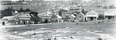 Black and white photograph of a row of business buildings on a rocky, sloping hillside, with a view across to residences and open land. Businesses from left to right (up the slope): J.E. Bishop, Coach Builders; Straughair Duncan, Engineers, Blacksmiths & Farriers; Straughair Duncan, Beechworth Foundry;  Wholesale & Retail Est. 1855 (?), Family Store Mackenzie; T. Pratten, Grocer - ?? & ?? Dealer, 