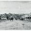 Uncropped black and white photograph of a man with moustache, dressed in hat and long riding coat and seated on a bob-tailed horse, stopped in the middle of a dirt road. He is outside a row of businesses, including R.M. Henzelsons Grain Store and Mackenzie Family Store (Wholesale & Retail Est. 1855), along a sloping hill. Behind him are a parked horse and laden two-wheeled cart backed up directly outside the granary verandah, and another man driving a horse and four-wheeled cart forward towards them. At the bottom of the hill is a bridge with forked road on the far side, leading to a number of houses on both sides of another hill. This is Bridge Road, Newtown, Beechworth.