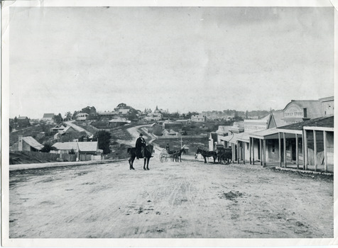 Uncropped black and white photograph of a man with moustache, dressed in hat and long riding coat and seated on a bob-tailed horse, stopped in the middle of a dirt road. He is outside a row of businesses, including R.M. Henzelsons Grain Store and Mackenzie Family Store (Wholesale & Retail Est. 1855), along a sloping hill. Behind him are a parked horse and laden two-wheeled cart backed up directly outside the granary verandah, and another man driving a horse and four-wheeled cart forward towards them. At the bottom of the hill is a bridge with forked road on the far side, leading to a number of houses on both sides of another hill. This is Bridge Road, Newtown, Beechworth.
