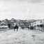 Black and white photograph of a man with moustache, dressed in hat and long riding coat and seated on a bob-tailed horse, stopped in the middle of a dirt road. He is outside a row of businesses, including R.M. Henzelsons Grain Store and Mackenzie Family Store (Wholesale & Retail Est. 1855), along a sloping hill. Behind him are a parked horse and laden two-wheeled cart backed up directly outside the granary verandah, and another man driving a horse and four-wheeled cart forward towards them. At the bottom of the hill is a bridge with forked road on the far side, leading to a number of houses on both sides of another hill. This is Bridge Road, Newtown, Beechworth.