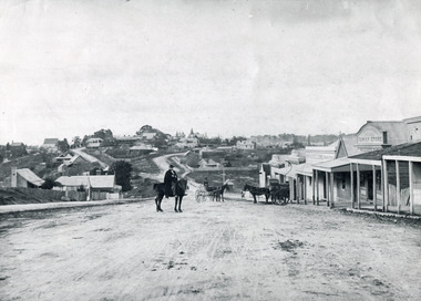 Black and white photograph of a man with moustache, dressed in hat and long riding coat and seated on a bob-tailed horse, stopped in the middle of a dirt road. He is outside a row of businesses, including R.M. Henzelsons Grain Store and Mackenzie Family Store (Wholesale & Retail Est. 1855), along a sloping hill. Behind him are a parked horse and laden two-wheeled cart backed up directly outside the granary verandah, and another man driving a horse and four-wheeled cart forward towards them. At the bottom of the hill is a bridge with forked road on the far side, leading to a number of houses on both sides of another hill. This is Bridge Road, Newtown, Beechworth.
