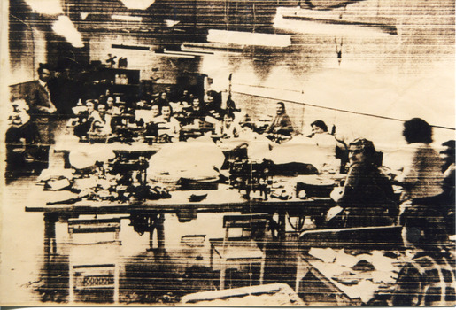 A busy workroom where women are seated at multiple long tables that have sewing machines and other equipment on them. There are approximately 15 women looking up from their work at the camera. One man stands to the left of the tables looking at the camera. One woman to the right of the tables appears in motion walking away from the camera.