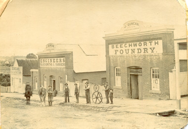 Six men and a boy stand outside of three buildings. The man furthest to the left is mounted on a horse, the next leftmost man holds the leadrope to another horse. The buildings have signs on them that read (from closest to furthest) "J. Duncan / Straughair and Duncan / Beechworth Foundry" "J. Duncan / Straughair and Duncan / Engineers Blacksmiths and Farriers" "Duncan's Coach Factory" 