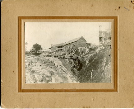 Two wooden buildings sit next to a waterfall with a mill wheel. Other buildings present in background, including a tall chimney