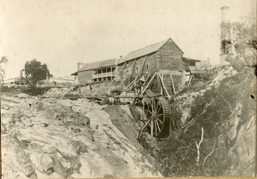 Two wooden buildings sit next to a waterfall with a mill wheel. Other buildings present in background, including a tall chimney.
