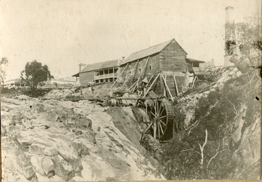 Two wooden buildings sit next to a waterfall with a mill wheel. Other buildings present in background, including a tall chimney.