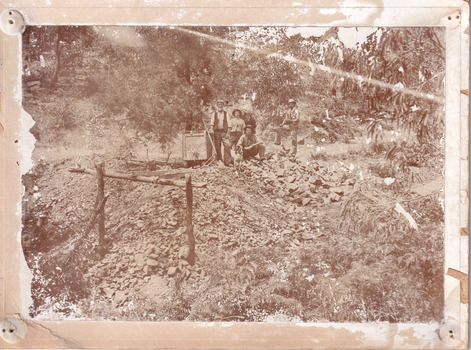 Four men and a dog standing at the entrance to 'Williams Good Luck Mine' (signposted on a wheeled cart on a track) in 1899. One man holds a second smaller dog. A large opening to a mine can be seen behind the men. Uneven rocks spill out of the mine entrance, with a timber frame in the foreground. A bush setting is around the mine. Image taken at the 800 ft Mopoke Gully-Stanley Tunnel.