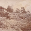 Four men relaxing under a wooden structure on four poles on a hilly landscape in 1899. One man stands nearby, in front of a stone wall. A dog is depicted standing by the four men, with one sitting man holding a smaller dog or puppy. Rock and dirt piles surround the men in an open bush setting. Some wooden logs can be seen stacked on top of each other in the middle of the photograph. 