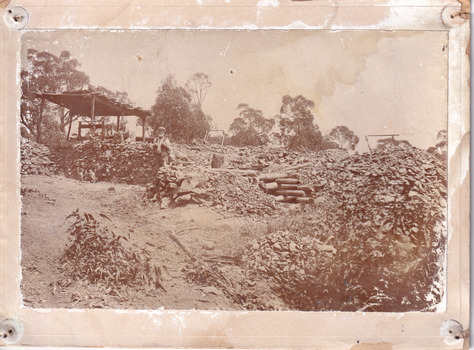 Four men relaxing under a wooden structure on four poles on a hilly landscape in 1899. One man stands nearby, in front of a stone wall. A dog is depicted standing by the four men, with one sitting man holding a smaller dog or puppy. Rock and dirt piles surround the men in an open bush setting. Some wooden logs can be seen stacked on top of each other in the middle of the photograph.