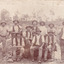 Sepia toned photograph of a group of 10 miners, some sitting on a log and some standing behind, with a dog in front of what appears to be an open cut mine.