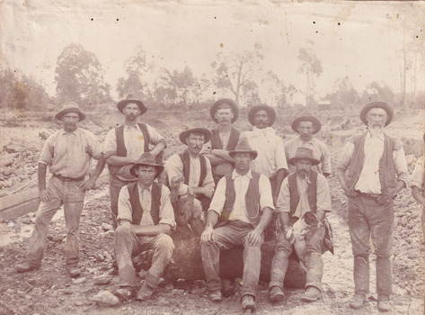 Sepia toned photograph of a group of 10 miners, some sitting on a log and some standing behind, with a dog in front of what appears to be an open cut mine.