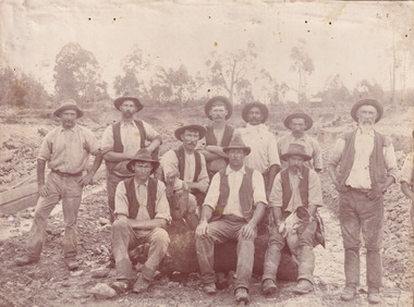 Sepia toned photograph of a group of 10 miners, some sitting on a log and some standing behind, with a dog in front of what appears to be an open cut mine.