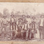 Sepia toned photograph of a group of 10 miners, some sitting on a log and some standing behind, with a dog in front of what appears to be an open cut mine.