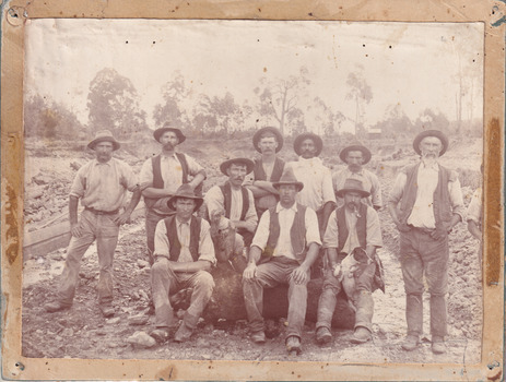 Sepia toned photograph of a group of 10 miners, some sitting on a log and some standing behind, with a dog in front of what appears to be an open cut mine.