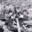 A sepia photograph of eleven miners from the Three Mile Miners standing and sitting around a plank of wood near a water race. In the background is a dirt wall.