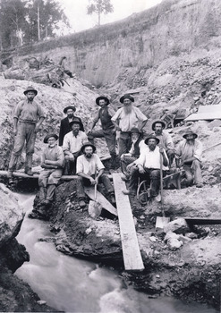 A sepia photograph of eleven miners from the Three Mile Miners standing and sitting around a plank of wood near a water race. In the background is a dirt wall.