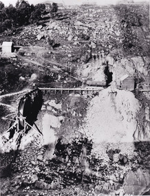 A black and white photograph taken from a high angle of rough terrain on the Rocky Mountain Mine. There is stone rubble spread across the landscape as well as mining equipment such as a vertical conveyor belt.