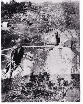 A black and white photograph taken from a high angle of rough terrain on the Rocky Mountain Mine. There is stone rubble spread across the landscape as well as mining equipment such as a vertical conveyor belt. There is a thin white border around the image.