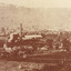 A landscape sepia photograph of a cleared landscape with very few trees and vegetation in the background. In the foreground, there is a clearing of land before a collection of wooden huts and tree stumps. Beyond this, the Rocky Mountain Mine can be seen in the distance with number of wooden buildings and a towering brick chimney. 