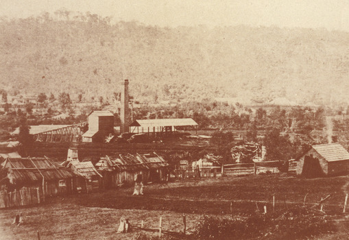 A landscape sepia photograph of a cleared landscape with very few trees and vegetation in the background. In the foreground, there is a clearing of land before a collection of wooden huts and tree stumps. Beyond this, the Rocky Mountain Mine can be seen in the distance with number of wooden buildings and a towering brick chimney. 