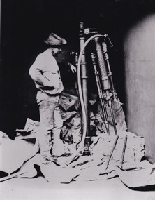 a man in a cowboy style hat (often worn in this era) standing next to mining machinery that is half a head taller than him