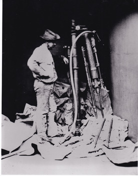 a man in a cowboy style hat (often worn in this era) standing next to mining machinery that is half a head taller than him