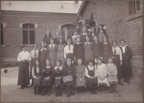 Photograph depicting a group of school girls and teachers posing outside a brick school building