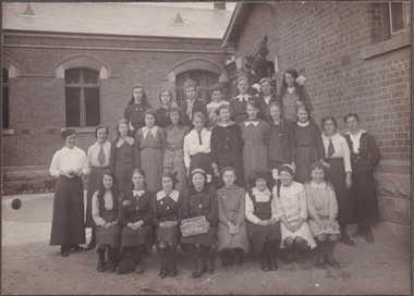 Photograph depicting a group of school girls and teachers posing outside a brick school building