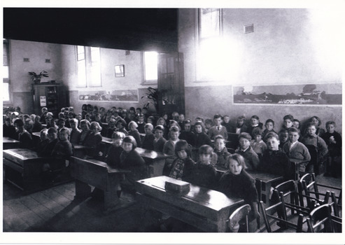Black and white photo of a crowded early 20th-century classroom with children seated at wooden desks.