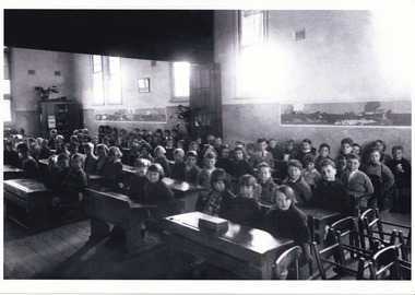 Black and white photo of a crowded early 20th-century classroom with children seated at wooden desks.
