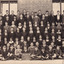 Black and white postcard depicting a large group of 79 students in formal attire in front of a brick building, one student in the middle front row holds a sign that reads "Beechworth H.E.S 1937".