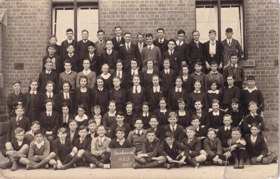 Black and white postcard depicting a large group of 79 students in formal attire in front of a brick building, one student in the middle front row holds a sign that reads "Beechworth H.E.S 1937".