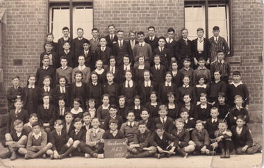 Black and white postcard depicting a large group of 79 students in formal attire in front of a brick building, one student in the middle front row holds a sign that reads "Beechworth H.E.S 1937".