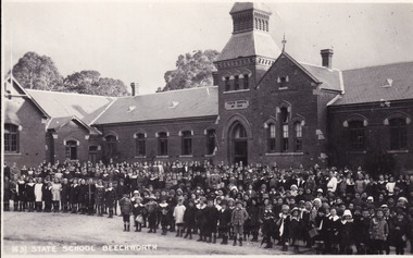 Black and white postcard of the Beechworth State School building. A large number of students and teachers standing in front of the building. 
