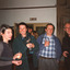 Group of two men and two women laughing and drinking in a room. Behind them are Indigenous artefacts mounted on the wall and a larger party of people.