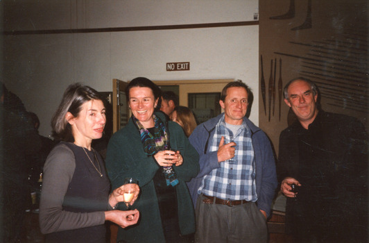 Group of two men and two women laughing and drinking in a room. Behind them are Indigenous artefacts mounted on the wall and a larger party of people.