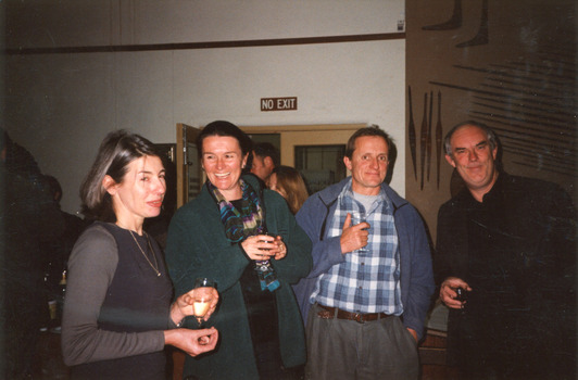 Group of two men and two women laughing and drinking in a room. Behind them are Indigenous artefacts mounted on the wall and a larger party of people.