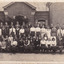 A photo of a group of school students, both boys and girls in uniforms, taking a group picture. There are 4 rows, the front two seated and the back two standing. In the background we can see an old style brick school building 