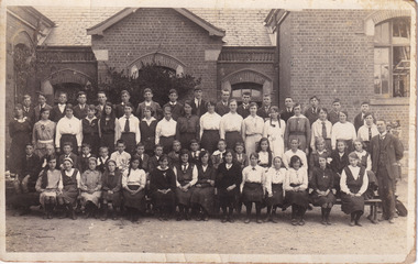 A photo of a group of school students, both boys and girls in uniforms, taking a group picture. There are 4 rows, the front two seated and the back two standing. In the background we can see an old style brick school building 
