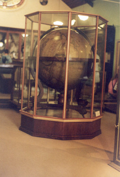 Large wooden globe of the world from the opposite perspective, encased in a large wood and glass display case. The photo is on a slight angle.