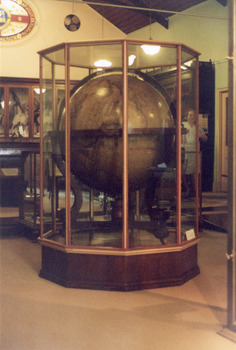 Large wooden world globe encased in a glass and wood display case, a person stands in the background behind the display case.