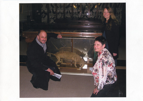 A crouching man and women surround a glass case containing a Thylacine, another women is standing next to the case. They are all smiling at the camera and behind them are hundreds of taxidermy specimens lining the wall, also in glass cases.