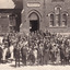 A sepia rectangular postcard with a large crowd of men and women standing in the foreground. Behind them is the Beechworth School building.
